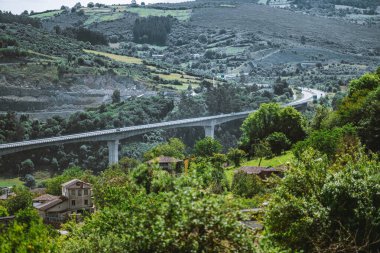Bereketli yeşil dağ vadisi boyunca uzanan uzun beton viyadük, güneşli bir günde uzak kayan tepeler, kırsal evler ve orman manzarasıyla telefoto merceğiyle görülüyor.