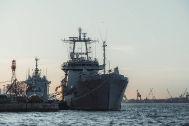 Gün batımında sanayi limanına demirlenmiş büyük deniz gemilerinin telefoto görüntüsü, vinçler ve sakin denizler dramatik deniz atmosferi ve güçlü çelik siluetler yaratıyor.