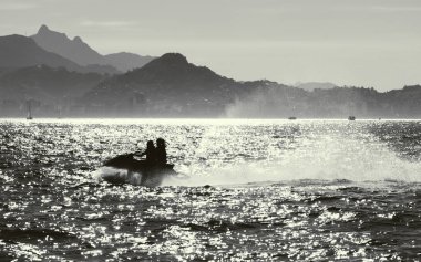 Arka planda sisli dağ manzarası olan, ışıldayan deniz suyu üzerinde kişisel bir su aracına binen iki insan silueti, parlak ışık yansıması, sinematik atmosfer yaratıyor.