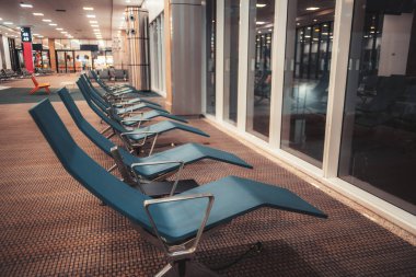 Modern mavi şezlonglu boş havaalanı terminali oturma alanı, büyük cam pencereler ve yapay ışıklandırma, biniş, seyahat ve ulaşım konseptinden önce sessiz bekleme alanı