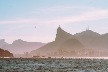 Rio de Janeiro 'nun telefoto deniz manzarası Corcovado' daki Kurtarıcı İsa, aşağıdaki sisli şehir ufuk çizgisi, Atlantik sularındaki yelkenliler ve pastel gökyüzündeki kuşlar.