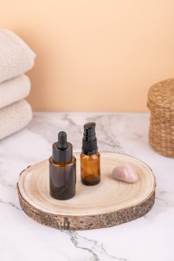 Two amber glass bottles with dropper and dispenser on wooden base with rose quartz, beige towels and wicker basket on marble table. Vertical composition with copy space. Concept of natural skincare and wellness lifestyle
