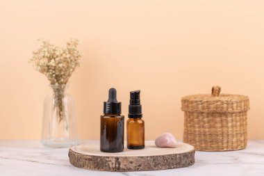 Front view of amber glass bottles with dropper and dispenser on wooden base with rose quartz stone, wicker basket and glass vase with dried flowers. Horizontal image with copy space. Concept of skincare and cozy lifestyle