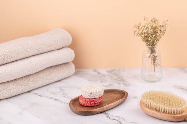 Solid shampoo bars on a wooden tray with a glass vase, three beige towels and a wooden brush. Peach background and marble surface. Minimalist vertical image with copy space. Concept of clean beauty and natural wellness