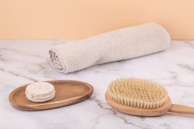 Solid shampoo bar on wooden tray with body brush and rolled towel on marble surface against peach background. Horizontal minimalist image with copy space. Concept of eco beauty and sustainable self care