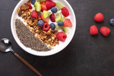 Kefir, granola, chia tohumu ve arkaplanda kaşık ve ahududulu taze meyve içeren beyaz bir kâse. Kopya uzayı ile yatay bileşim. Sağlıklı kahvaltı ve temiz yemek kavramı