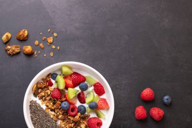 Kefir, kırmızı meyve, kivi ve chia ile sağlıklı bir kahvaltıyı koyu arka planda, dağınık granola ve ahududu ile birlikte beyaz bir kasede izlemenin en güzel görüntüsü. Kopya uzayı ile yatay çekim. Doğal beslenme ve refah kavramı