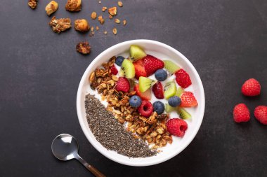 Kefir, chia, granola ve karışık meyveli beyaz kase koyu yüzeyde dağınık granola, ahududu ve bir kaşık ile. Kopyalama alanı olan en düşük stil. Dengeli beslenme ve sabah rutini kavramı