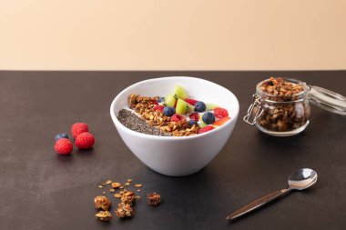 Kefir, chia tohumu, granola ve kivi, ahududu ve yabanmersini gibi taze meyvelerle bej arka planda ve dağınık granola ve ahududu içeren koyu renkli bir yüzey. Uzayı kopyala Sağlıklı kahvaltı kavramı ve