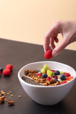 Kadın eli beyaz bir kaseye kefir, chia, granola ve bej arka planda taze meyvelerle ahududu yerleştiriyor. Kopya uzayı ile dikey bileşim. Temiz yemek ve sağlık yaşam tarzı kavramı