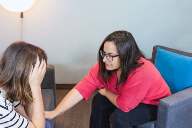 Gözlük takan yetişkin bir kadın ve fuşya süveteri sıcacık ve minimalist dekorlu rahat bir terapi odasında sıkıntılı genç bir kadını rahatlatıyor. Boşluğu kopyala