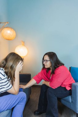 Terapi seansı sırasında endişeli genç bir kadını teselli eden kadın psikolog, empatik fiziksel temas ve anlayış ifadesi. Dekoratif lambalı açık mavi arkaplan. Uzayı kopyala Psikolojik yardım ve akıl sağlığı kavramı