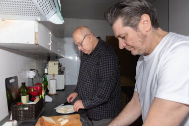 İki yetişkin adam bir ev mutfağında birlikte aperatifler hazırlıyor, sıcak, gerçek ve günlük atmosfer, fotokopi alanı ile yatay oryantasyon. Dostluk kavramı ve günlük rutin