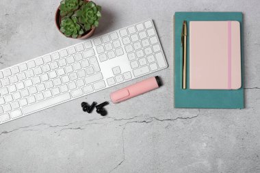 Beyaz klavyesi, pembe defteri, kalemi, kablosuz kulaklığı ve gri taş arka planda sulu bitkisi olan modern çalışma alanının üst görünümü. Uzayı kopyala Üretkenlik kavramı, uzak çalışma ve ofis yaşam tarzı