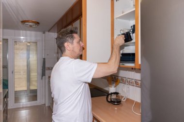 Ev mutfağında kahve hazırlarken dolaptan bardak alan gerçek bir yetişkinin yan görüntüsü. Gerçek bir ev ortamında otantik günlük yaşam tarzı sahnesi. Gerçek hayat kavramı, günlük rutin ve ev hayatı.