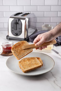 Erkek el, ahşap maşayla taze yapılmış tost ekmeğini tutuyor. Daha fazla tost makinesi, reçel kavanozu, tereyağı, fincan ve peçeteli gri tabak. Basit yemek ve konforlu ev kavramı