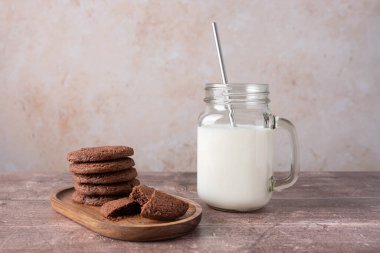 Bir kırık kurabiye ve bir kavanoz süt, bej taştan arka plan ve ahşap masa, fotokopi alanı olan yatay kompozisyon içeren ahşap bir tepside çikolatalı kurabiyeler. Sıcak tatlı ve konforlu yemek kavramı