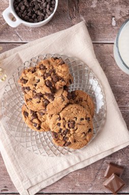 Bir bardak süt ve çikolata parçalarıyla bej kumaş ve ahşap masa üzerindeki cam tabakta çikolatalı kurabiyelerin üst görüntüsü. Ev yapımı tatlı ve sıcak yemek kavramı
