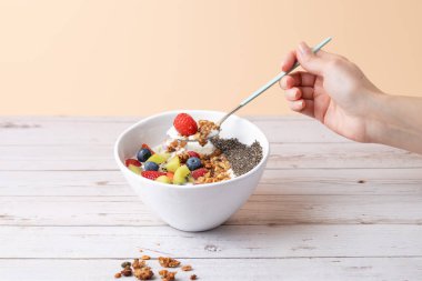 Kadın eli kahvaltı kasesinden bir kaşık kefir, meyve, chia tohumu ve granola alıyor. Pastel arka planda dengeli bir kompozisyon. Uzayı kopyala Sağlıklı kahvaltı ve günlük an kavramı