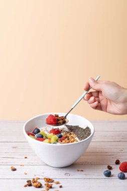 Elinde bir kaşık granola ve kefir, taze meyve ve chia tohumlarıyla beyaz bir kasenin üzerinde ahududu tutan bir kadın eli. Pastel arka plan ve kırsal yüzey. Uzayı kopyala Sağlıklı rutin ve dikkatli beslenme kavramı
