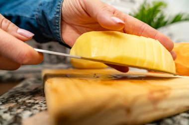 Kadın taze malzemeler hazırlıyor, tahta bir tahtanın üzerinde soyulmuş bir patates kesiyor.