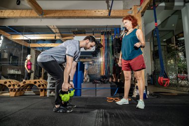 Kadın antrenör spor salonunda onu izlerken erkek ağırlık egzersizi yapıyor.