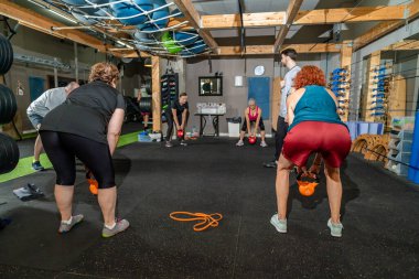 Bir grup yetişkin spor salonunda bir antrenörle kettlebell salıncakları yapıyor.