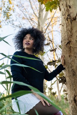 Sonbaharda dışarıda poz veren siyah kadın, düşünceli bir ifadeyle yukarı bakıyor.