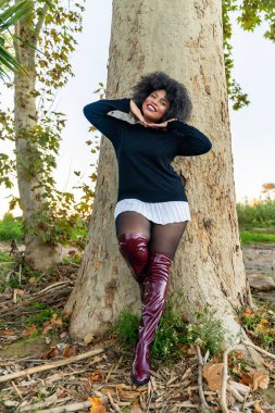 Gülümseyen kadın ağaca yaslanıyor siyah bir kazak ve diz üstü çizmeler giyiyor.