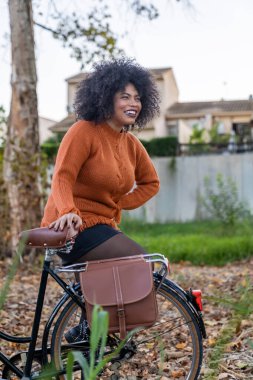 Kıvırcık saçlı, gülen kadın dışarıda sonbahar gezintisi sırasında bisiklette duruyor.