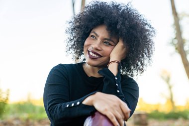 Genç siyahi kadın gülümsüyor, düşünceli ve mutlu bir ifadeyle başka tarafa bakıyor.