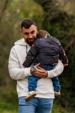 Genç bir baba, dışarıda bir çocuğu taşıyor ve teselli ediyor. Aile sevgisini ve bakımını simgeliyor.