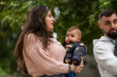 Bir kadın kucağında bebek tutarken bir adam yakınlarda duruyor ve bir aile sahnesi yaratıyor.