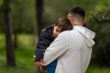 Anne ve baba, açık hava ortamında, omzunun üzerinden korunarak uyuyan küçük bir çocuğu taşıyor.