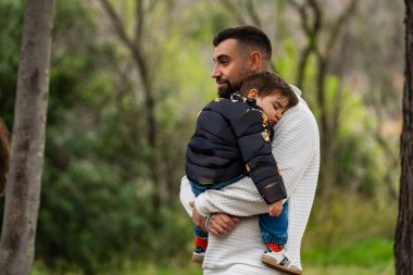 Park ortamında çocuğunu kucağında sakince uyuyan bir adam.