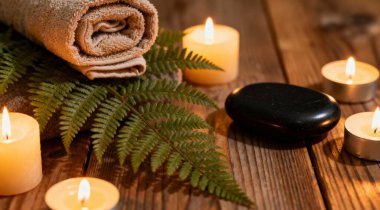 Eğreltiotlu havlular, mumlar ve ahşap arka planda siyah sıcak taşlar. Mumlarla aydınlatılmış sıcak taş masaj seti.