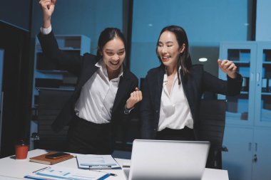 İki Asyalı iş kadını şirket stratejisini tartışıyorlar. Gece ofiste dizüstü bilgisayar ve grafikler kullanıyorlar.