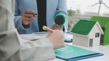 Masasında bir model ev ve rüzgar türbini olan sürdürülebilir ve çevre dostu gayrimenkul projelerini tartışan profesyoneller.