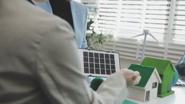 Masasında bir model ev ve rüzgar türbini olan sürdürülebilir ve çevre dostu gayrimenkul projelerini tartışan profesyoneller.