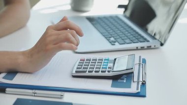 İki finans muhasebecisi dizüstü bilgisayardaki iş tablolarını analiz ediyor, ofis masasında oturuyor, kağıt belgelerine bakıyor, veri tabanlarıyla çalışan hesap makinesi kullanıyor, üstün tabloları kontrol ediyor.