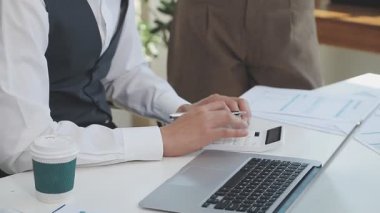 İki finans muhasebecisi dizüstü bilgisayardaki iş tablolarını analiz ediyor, ofis masasında oturuyor, kağıt belgelerine bakıyor, veri tabanlarıyla çalışan hesap makinesi kullanıyor, üstün tabloları kontrol ediyor.