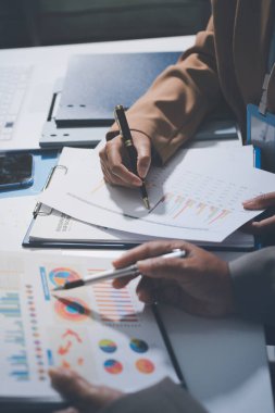 İş arkadaşları, modern bir ofis ortamında yapılan önemli bir toplantıda strateji ve piyasa performansı hakkında tartışırken finansal tablolar ve raporları analiz ediyorlar.