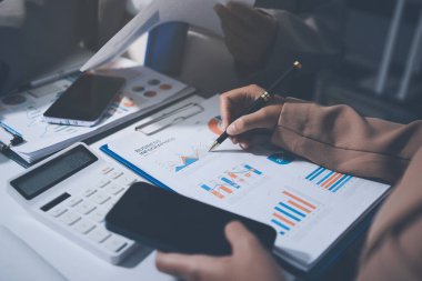 İş arkadaşları, modern bir ofis ortamında yapılan önemli bir toplantıda strateji ve piyasa performansı hakkında tartışırken finansal tablolar ve raporları analiz ediyorlar.