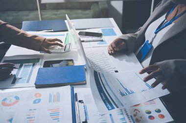 İş arkadaşları, modern bir ofis ortamında yapılan önemli bir toplantıda strateji ve piyasa performansı hakkında tartışırken finansal tablolar ve raporları analiz ediyorlar.