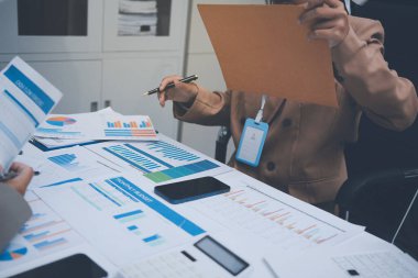 İş arkadaşları, modern bir ofis ortamında yapılan önemli bir toplantıda strateji ve piyasa performansı hakkında tartışırken finansal tablolar ve raporları analiz ediyorlar.