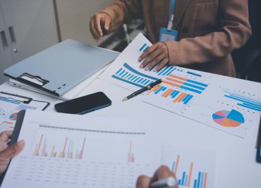 İş arkadaşları, modern bir ofis ortamında yapılan önemli bir toplantıda strateji ve piyasa performansı hakkında tartışırken finansal tablolar ve raporları analiz ediyorlar.