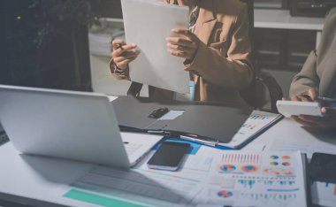 İş arkadaşları, modern bir ofis ortamında yapılan önemli bir toplantıda strateji ve piyasa performansı hakkında tartışırken finansal tablolar ve raporları analiz ediyorlar.