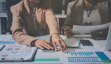 İş arkadaşları, modern bir ofis ortamında yapılan önemli bir toplantıda strateji ve piyasa performansı hakkında tartışırken finansal tablolar ve raporları analiz ediyorlar.