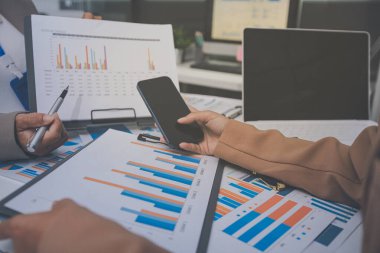 İş arkadaşları, modern bir ofis ortamında yapılan önemli bir toplantıda strateji ve piyasa performansı hakkında tartışırken finansal tablolar ve raporları analiz ediyorlar.