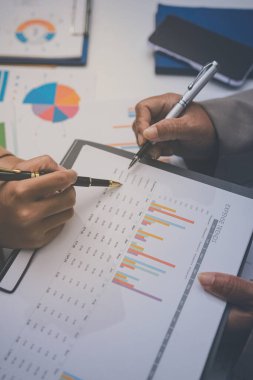 İş arkadaşları, modern bir ofis ortamında yapılan önemli bir toplantıda strateji ve piyasa performansı hakkında tartışırken finansal tablolar ve raporları analiz ediyorlar.